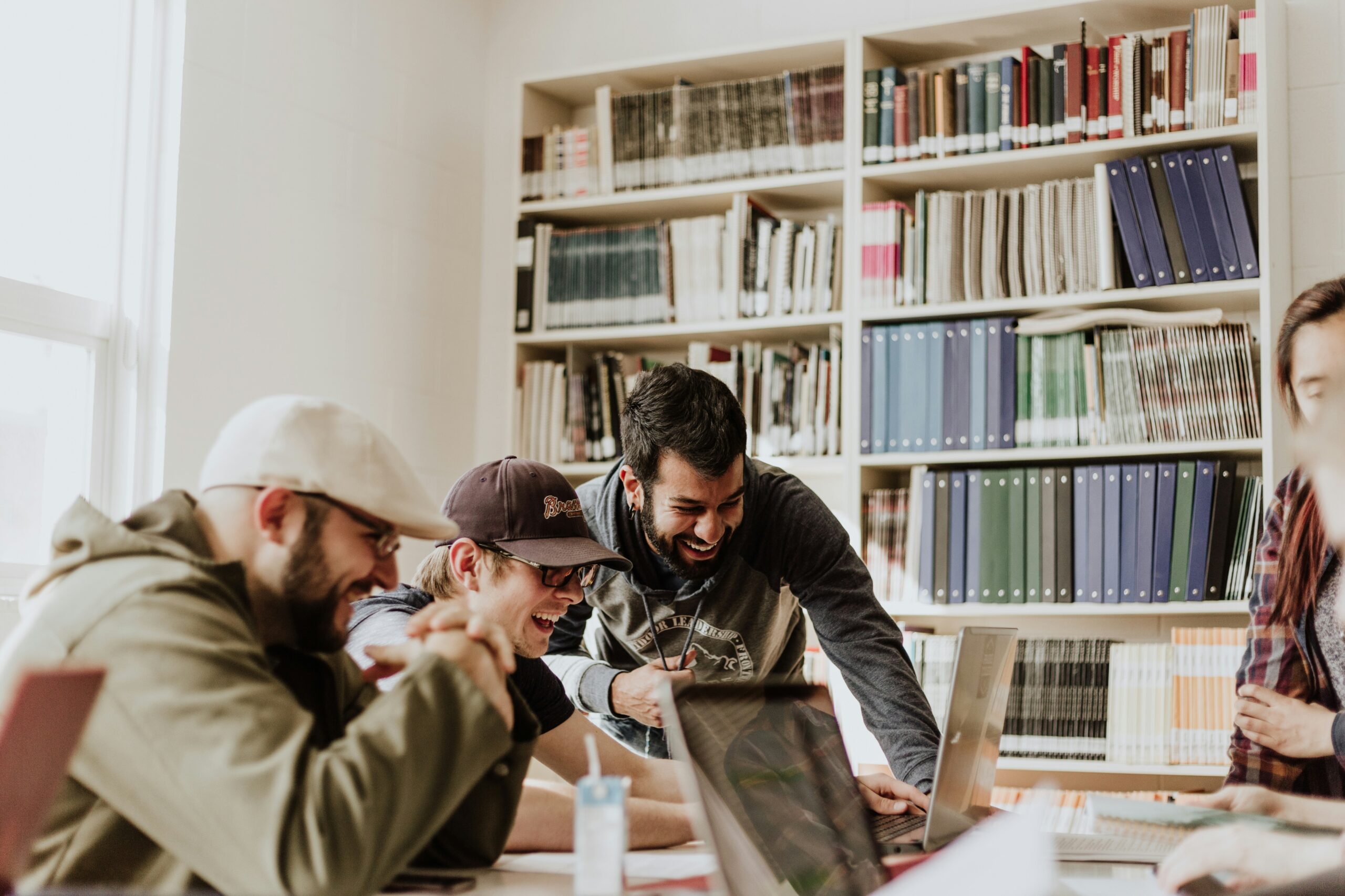 Group of young adults laughing and collaborating around a laptop in a library or study setting. The atmosphere is friendly and engaging, with everyone focused on the shared task, conveying teamwork, learning, and positive interaction.